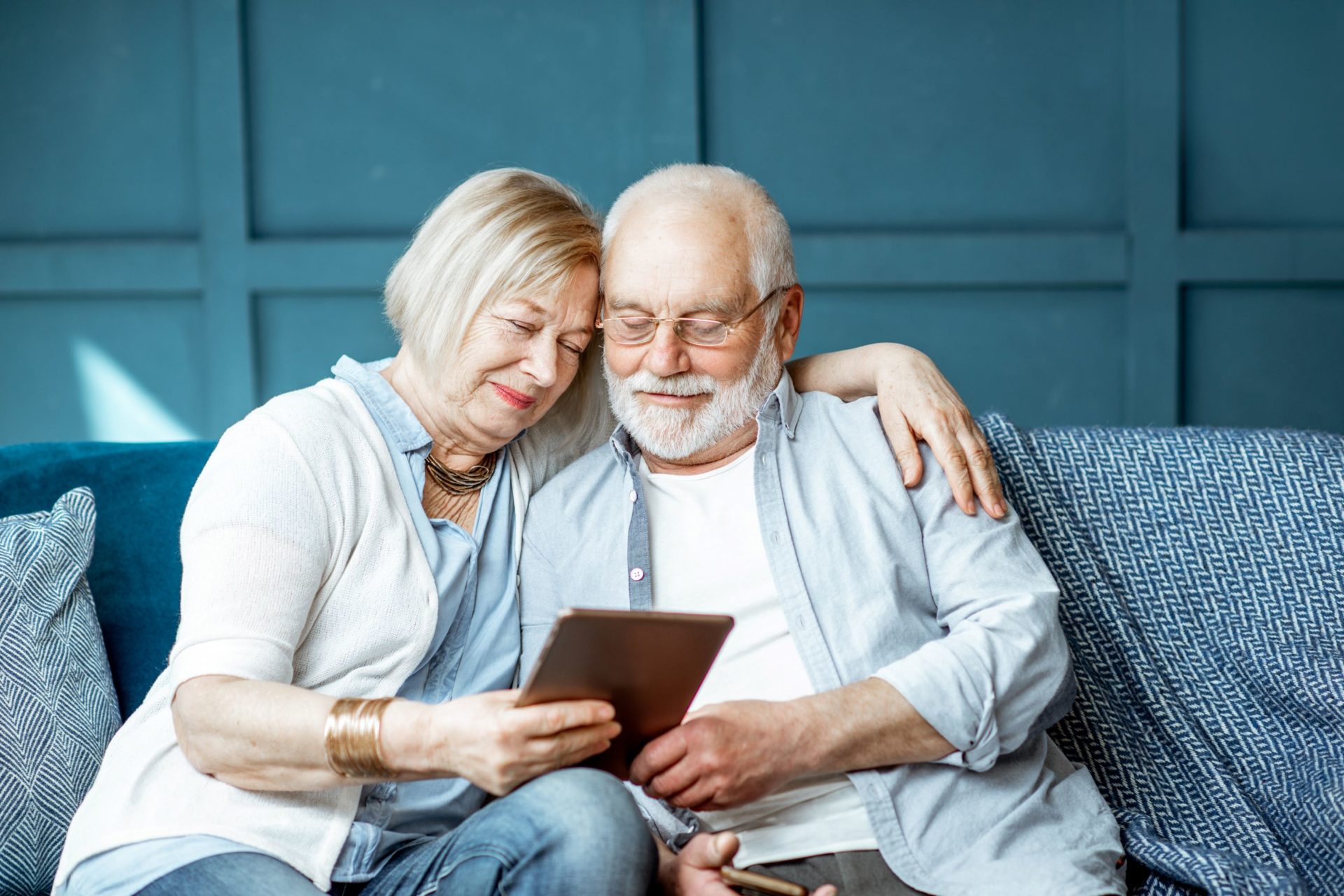 An old couple sat together on a sofa looking down at their iPad.