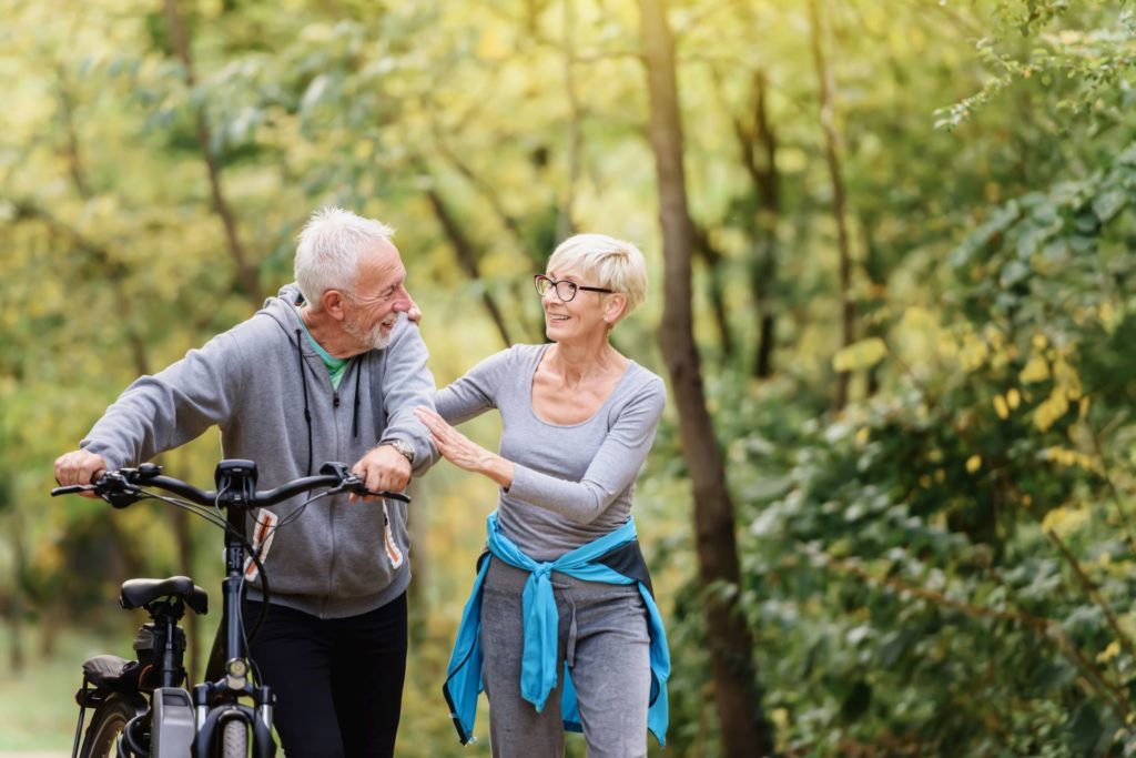 a couple walking through a forest, the man is walking with his bike as the woman and him talk.